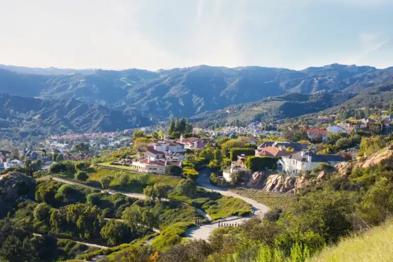 A scenic view of Pacific Palisades, California, showing winding roads, hillside homes, lush greenery, and distant mountain ranges under a clear sky.