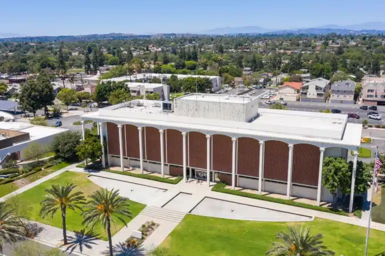 Daytime aerial view of Fullerton City Hall in California, showcasing its mid-century modern architecture with tall columns, manicured lawn, palm trees, and surrounding suburban neighborhood.