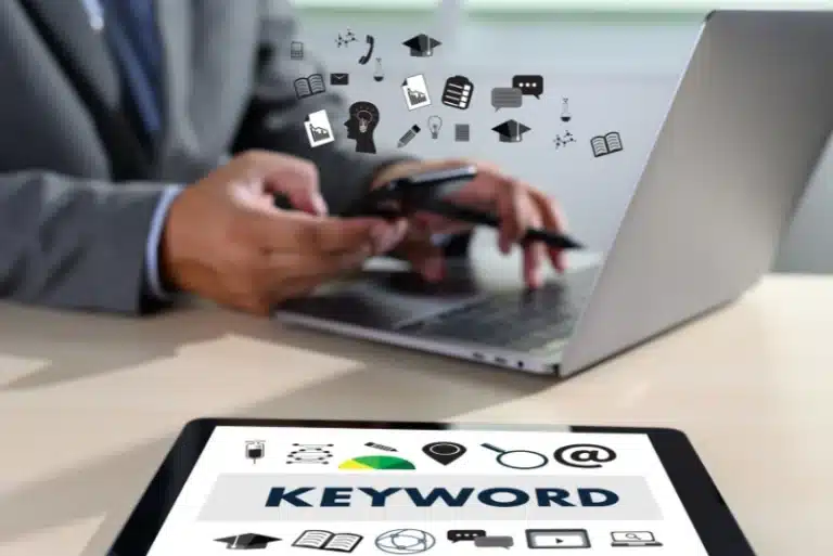 Person using a smartphone and laptop with digital marketing icons floating above, and a tablet in the foreground displaying the word “KEYWORD